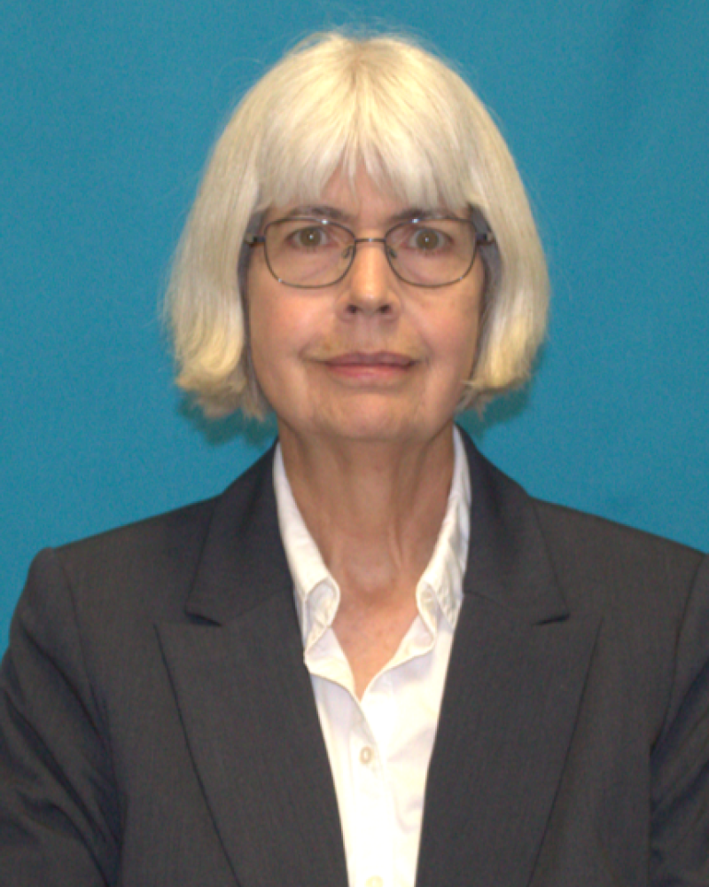 Photograph of a person with shoulder-length blonde hair and glasses wearing a dark suit jacket over a white collared shirt, set against a solid blue background. 