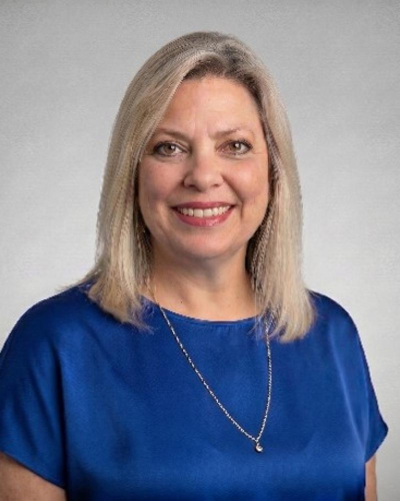 Photograph of a person who is smiling, with shoulder-length blonde hair, wearing a blue blouse and a silver necklace, standing against a plain gray background. 